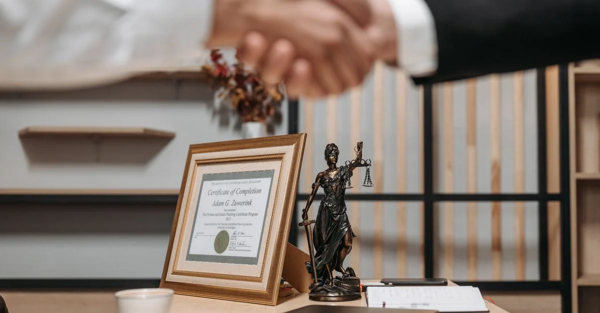 Une poignée de main professionnelle dans un bureau avec un diplôme et une statue de la Justice, symbolisant le droit et la réussite.