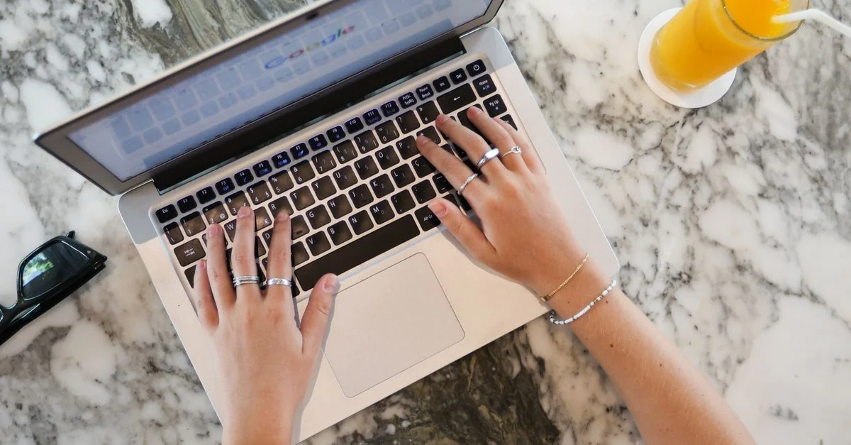 Des mains tapant sur un ordinateur portable avec un verre de jus d'orange sur une surface en marbre, idéal pour des thèmes de télétravail.